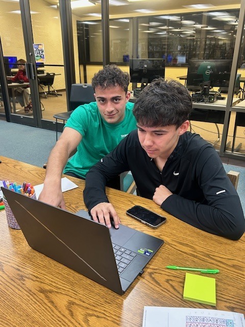 Two students collaborate on laptop at table with notebooks and pens nearby.