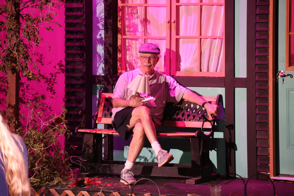 Older man sits on bench under pink lighting holding prop and smiling.