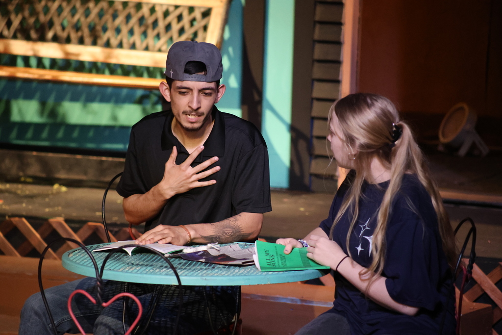 Two actors seated at small table reading scripts during rehearsal scene.