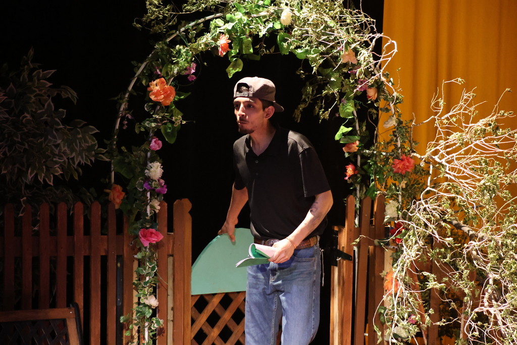 Man enters garden set through floral arch holding script pages on stage.