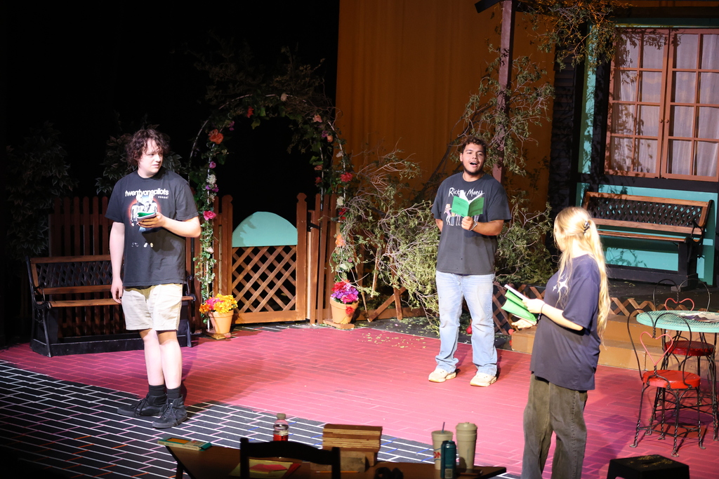 Three actors spread across stage performing with garden set and props.
