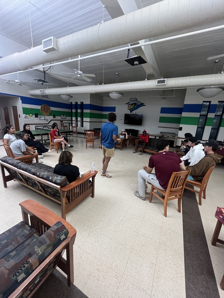 People sit in chairs around a large room playing a game with a man standing in the center.