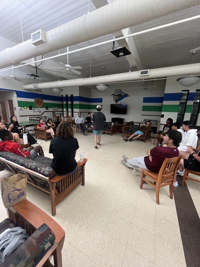 People sit in chairs around a large room playing a game with a man standing in the center.