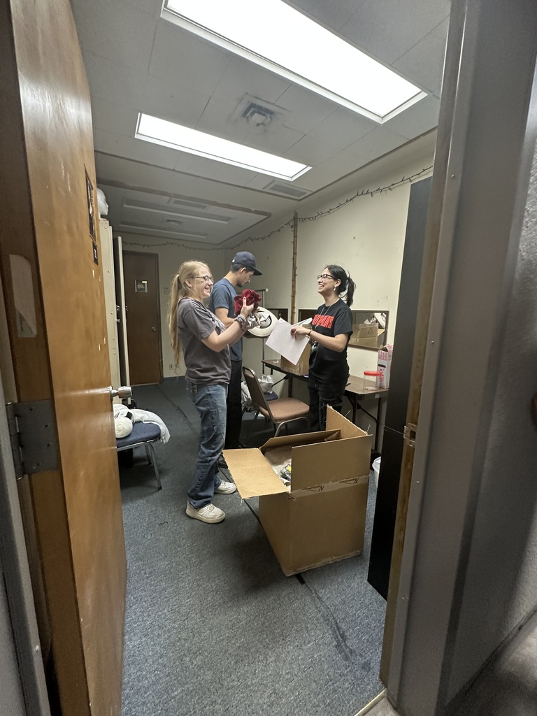 Three students laugh while organizing costume pieces and paperwork backstage