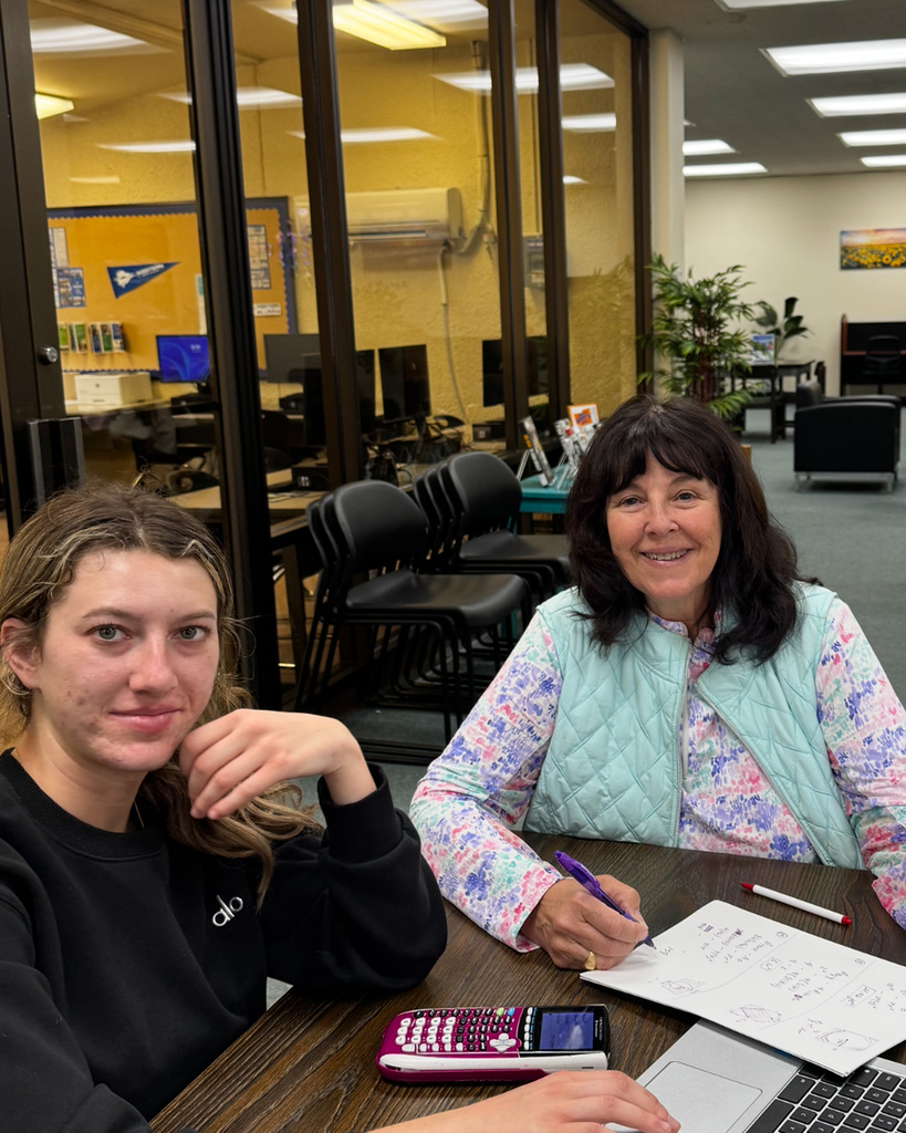 Ms. Perry writes in a notebook while a student types on their laptop, both smiling for the camera.