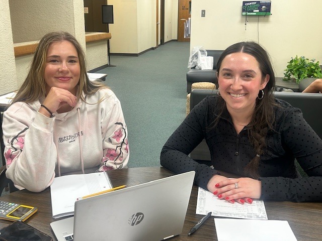 Brianna Ward (right) and a student women smile for the camera sitting at a table in the Library.