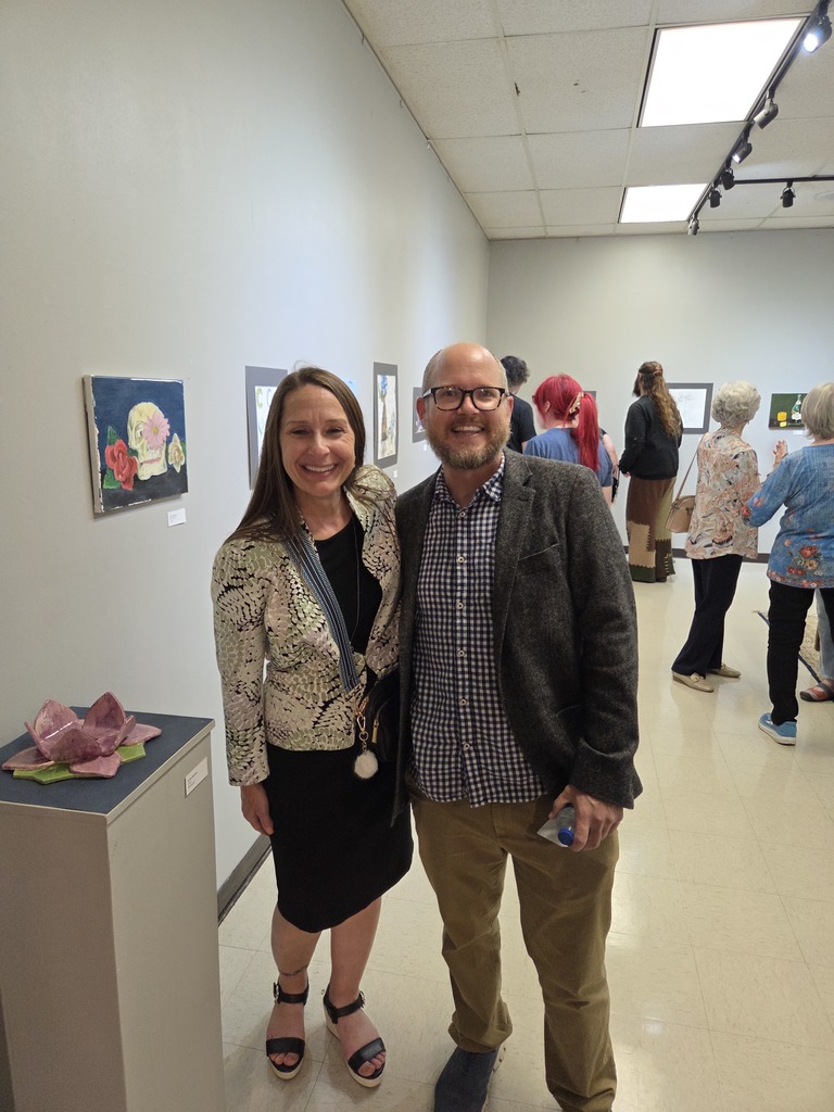 Dr. Sharp and Patrick Schneider  smile in gallery surrounded by student artwork displays.