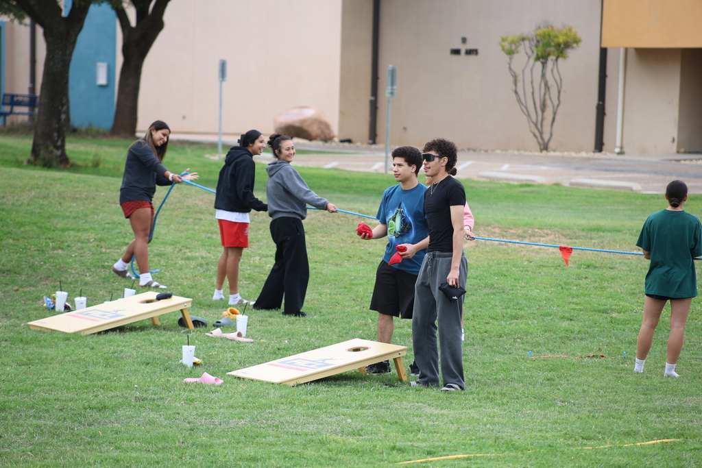 Students prepare for tug-of-war beside cornhole boards on campus lawn.