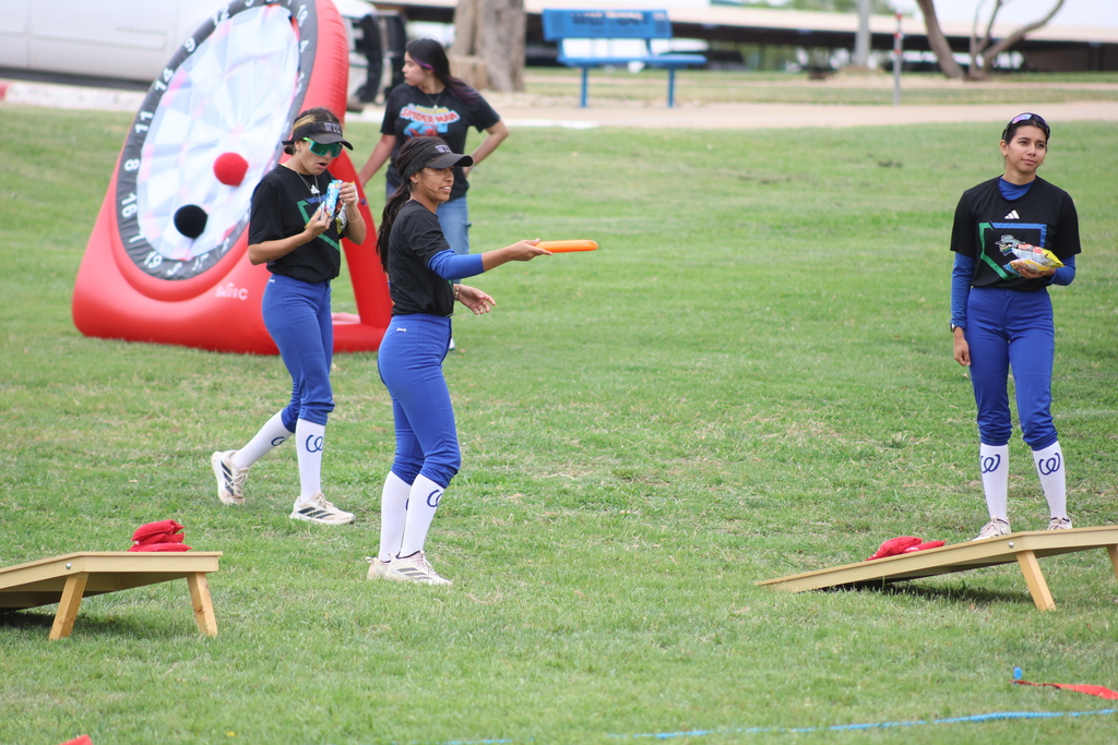 Softball players toss frisbee near cornhole boards and inflatable target.