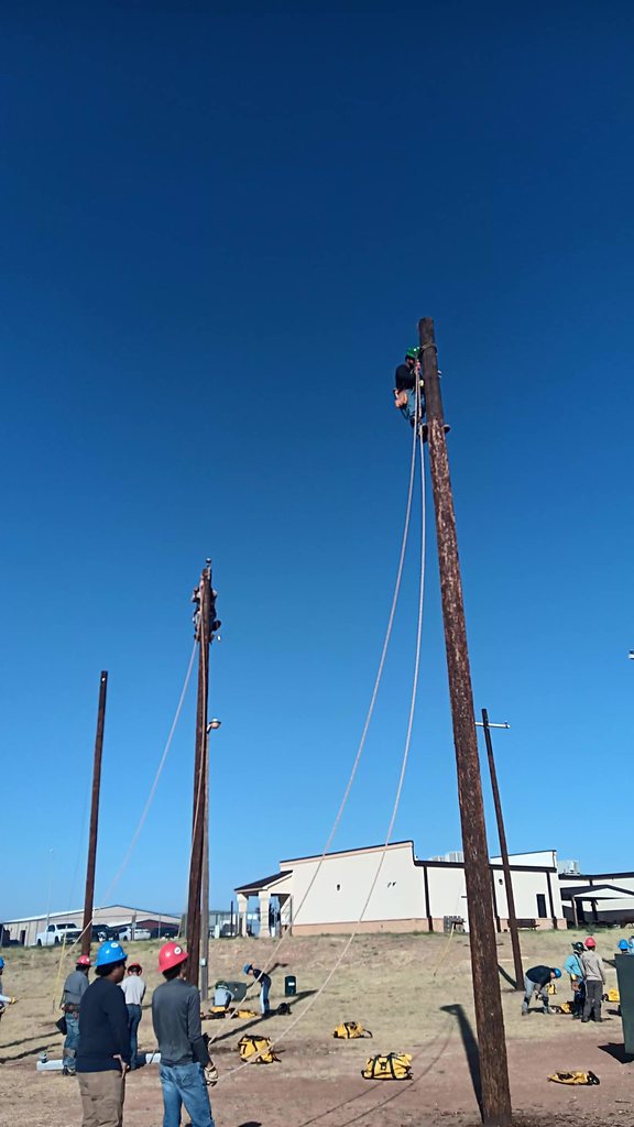 An electrical lineworker student climbs to the top of a pole as students on the ground watch.