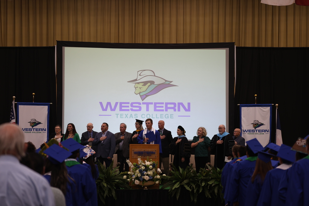 Graduate speaks at podium during ceremony while faculty and administrators stand behind on stage.