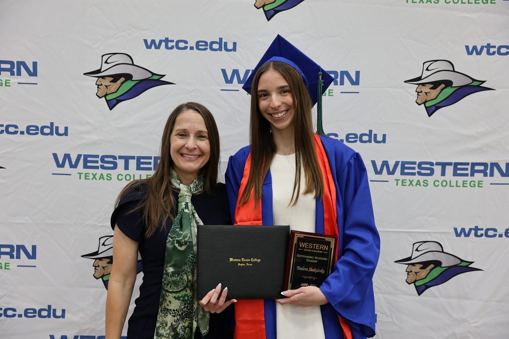 Graduate in blue cap and gown holds diploma and award plaque while posing with woman in front of WTC backdrop.