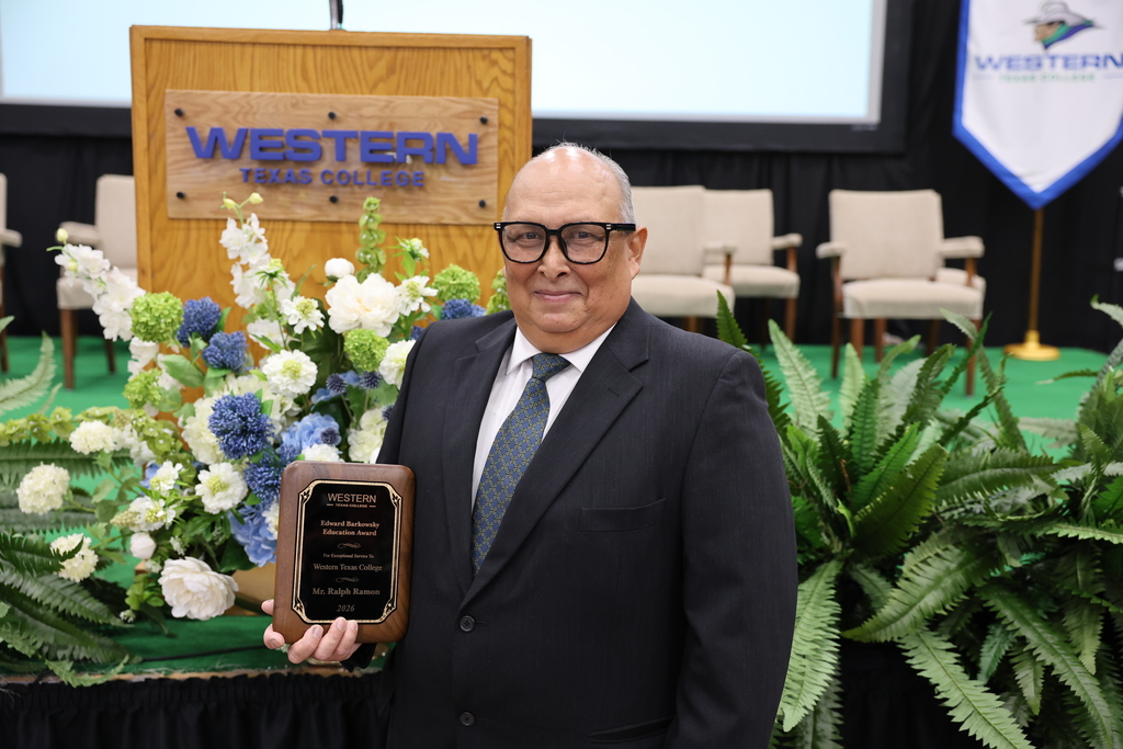 Man holds Western Texas College award plaque while standing in front of stage decorations.