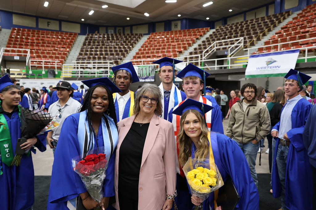 Group of graduates in blue caps and gowns pose with woman, holding bouquets, inside arena after commencement ceremony.