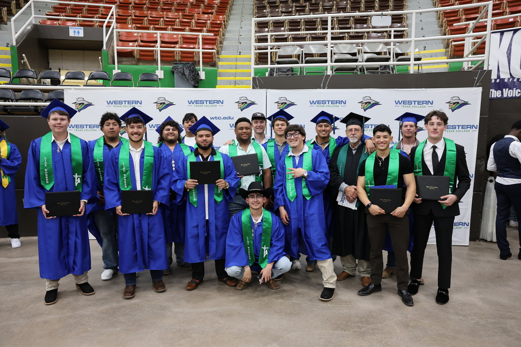 Group of graduates in blue gowns and green stoles pose together holding diplomas and smiling.