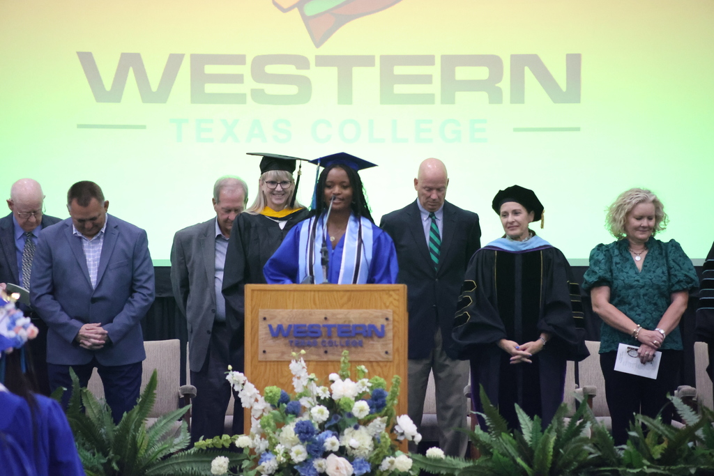 Graduate stands at podium during ceremony with faculty seated behind and Western Texas College logo projected.