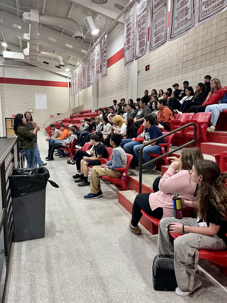 Two women speak to students sitting in seats in a gym.