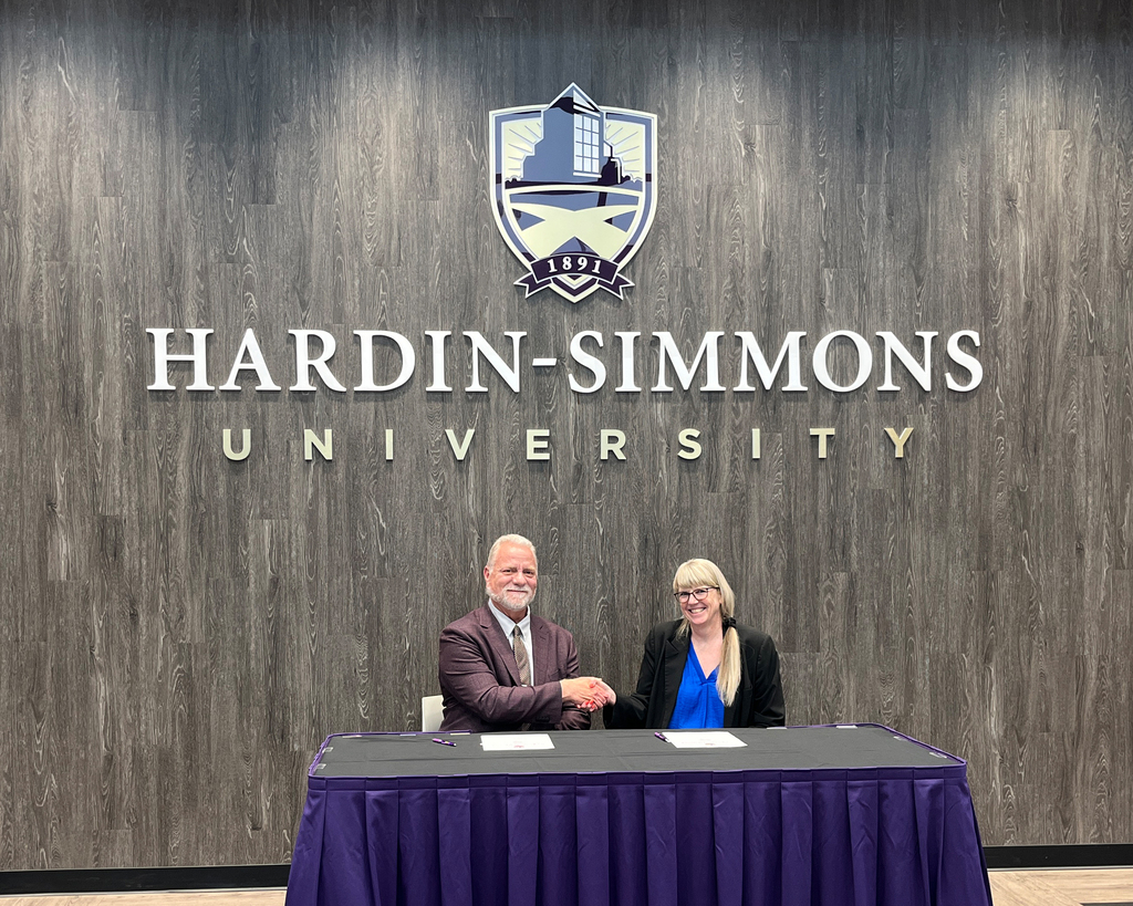 Dr. McNair and Stephanie Ducheneaux shake hands sitting at a covered table with the Hardin-Simmons University logo on the wall behind them.