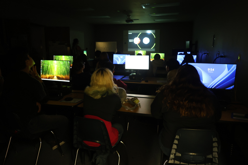 Students watch educational presentation in computer lab with monitors illuminated in darkened room.