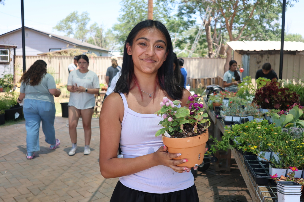 A student smiles while holding a potted flowering plant in an outdoor garden nursery, with other students participating in gardening activities in the background.