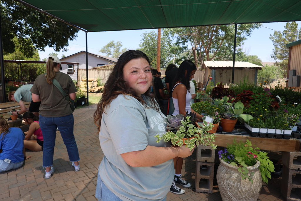 A student holds a tray of succulents while standing beside a table of plants as other students participate in a gardening activity under a shaded outdoor workspace.