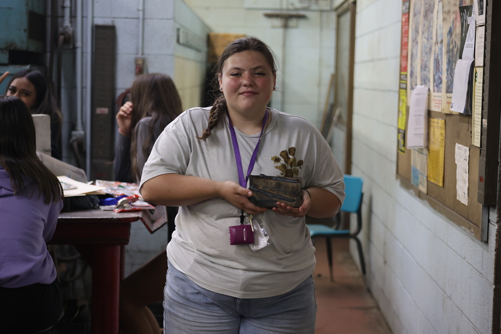 A student holds a piece they made while standing in an welding shop, with classmates working on projects in the background.