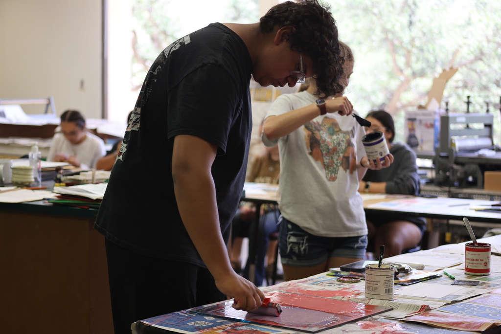 A student uses a brayer to apply ink to a printing plate while classmates prepare materials and work on printmaking projects in an art studio.