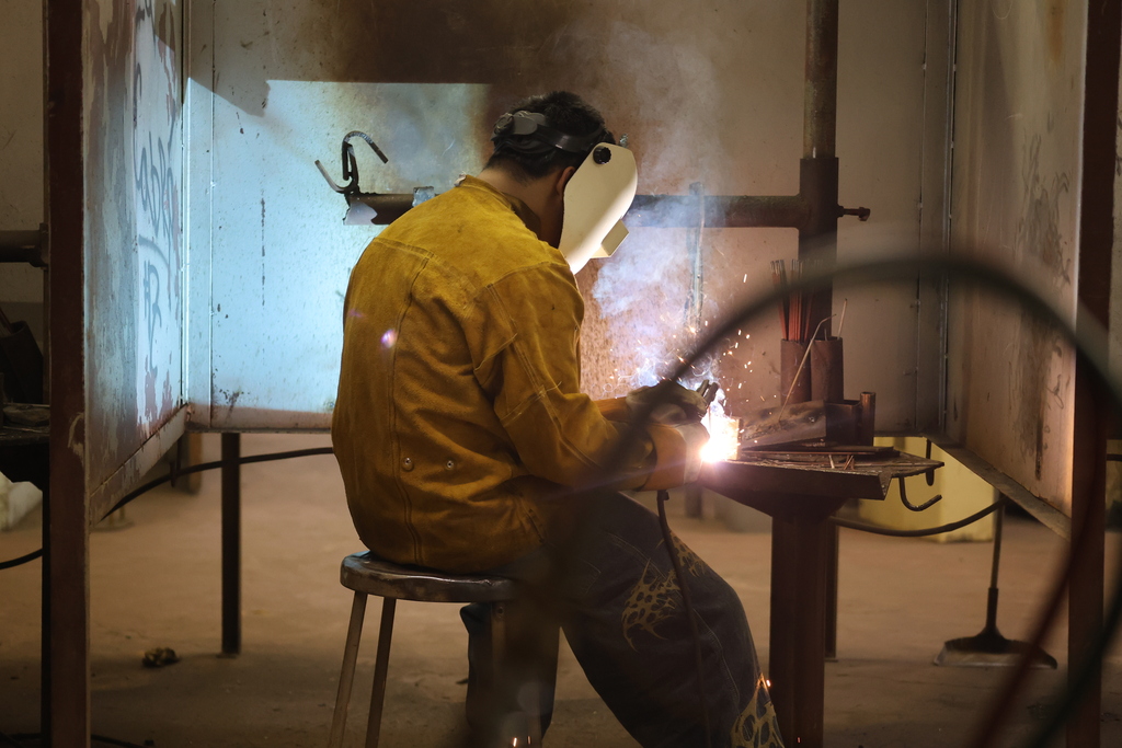 A student wearing welding safety gear welds metal inside a welding booth as sparks and light illuminate the workspace.