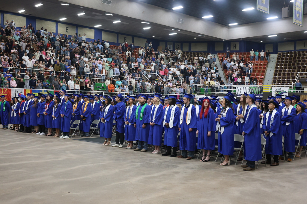 WTC graduates wearing graduation gowns stand in front of their chairs at commencement.