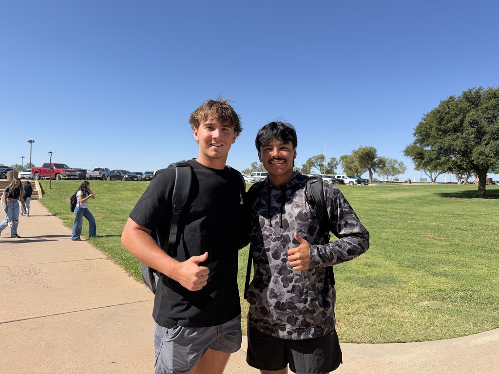 Two WTC students wearing backpacks smile and give thumbs-up gestures outside on campus.