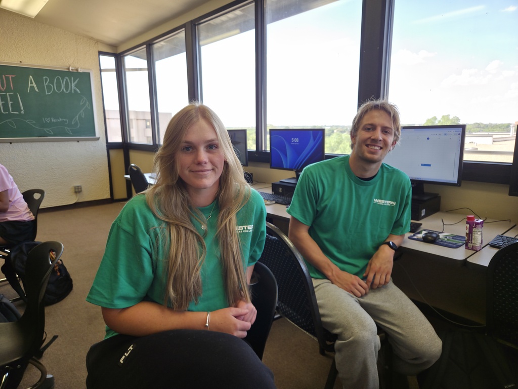 Two Western Texas College student tutors wearing green WTC shirts sit together in a campus computer lab.