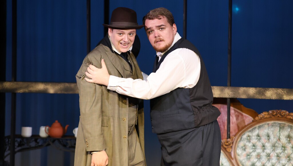 Two actors in period costumes perform a scene from the play on stage.