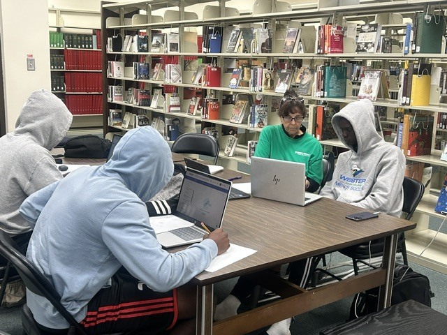 A tutor helps a student while other students study at a table.