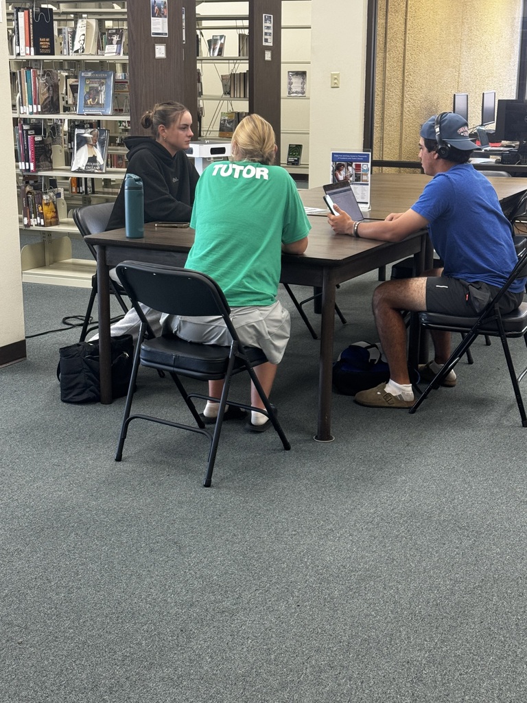 A tutor in a tutoring t-shirt sits a a table helping two students.