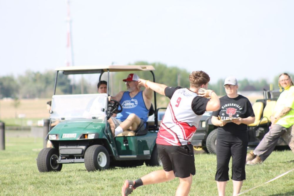Male athlete preparing to throw shot put while a woman in black holds a clipboard and watches, a green golf cart is in the background with two passengers and a spectator in bright yellow shirt watches from the right side of the background