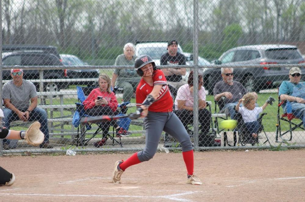 Female athlete in red uniform shirt and grey uniform pants with tall red socks swinging a bat at an incoming yellow softball while spectators watch on in the background
