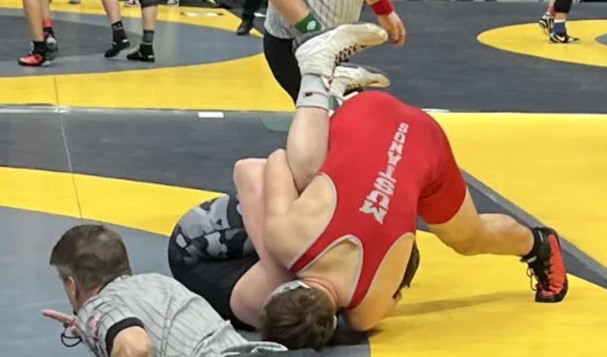 A male wrestler wrestling an opponent in black and grey camouflaged singlet on a yellow and grey mat with a referee in the bottom left corner blowing a whistle