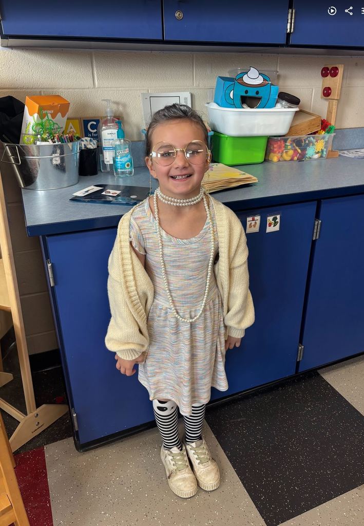 A young girl smiling in front of blue cabinets, dressed as an elderly woman with grey hair, oversized round glasses, a pearl necklace, and a cardigan.