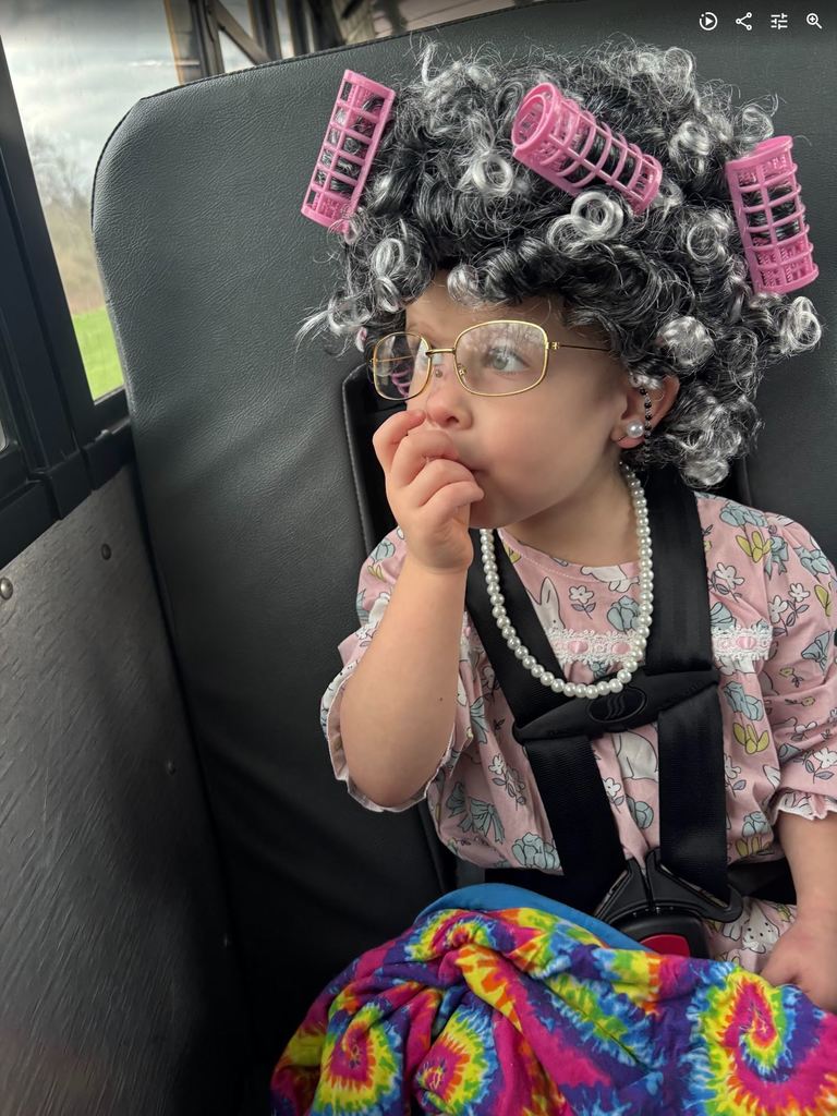 A young girl sitting on a bus wearing a grey wig with pink hair rollers, gold-rimmed glasses, and a pearl necklace.