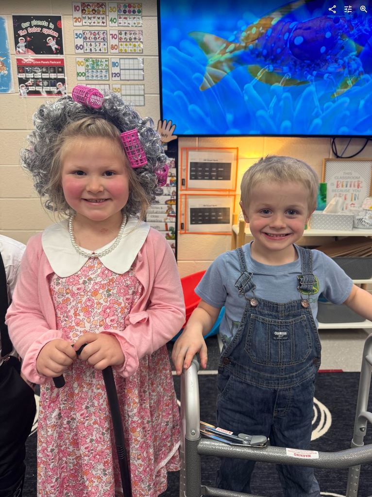 Two preschoolers posing together; one girl in a floral dress with a grey wig and a cane, and one boy in denim overalls leaning on a child-sized walker.