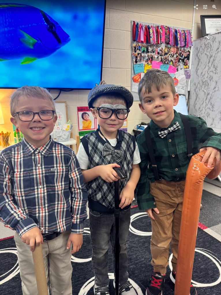 Three preschool boys posing together in "old man" costumes, featuring flat caps, suspenders, bow ties, drawn-on grey eyebrows, and walking canes.