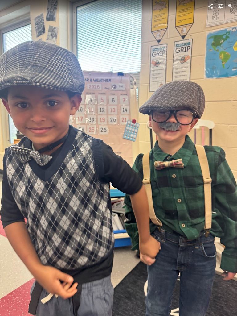 Two young boys holding hands and smiling, dressed in flat caps, sweater vests, and plaid shirts, with one wearing a grey costume mustache.