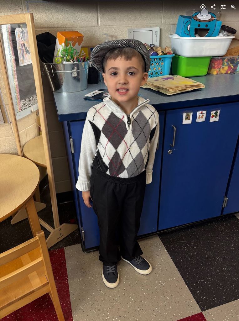 A preschool boy smiling in a classroom, dressed as an older man wearing a grey flat cap, an argyle sweater vest, and black slacks.