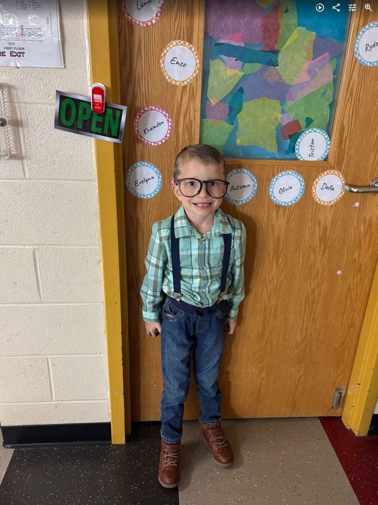 A preschool boy standing by a classroom door wearing oversized black glasses, blue suspenders, and a plaid shirt.