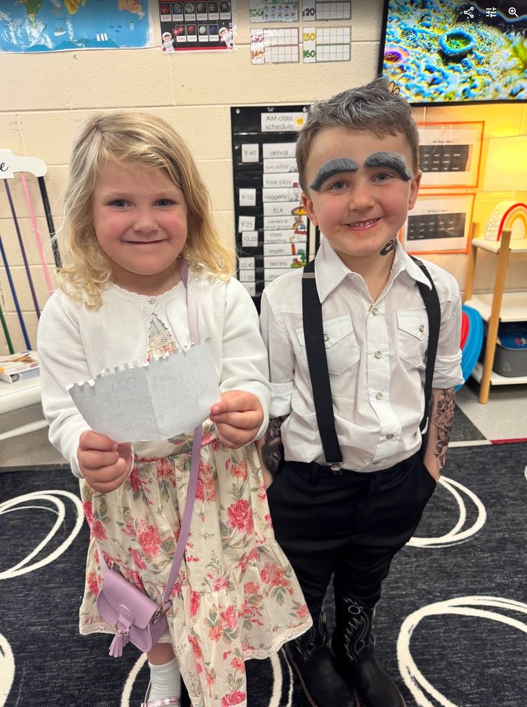 A young girl in a floral dress and a boy with thick grey drawn-on eyebrows, suspenders, and "tattoo" arm sleeves posing together.