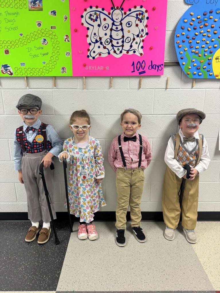 Four young children stand in a line against a white cinderblock wall, dressed up as elderly people for "100 Days of School." They wear various costumes including flat caps, suspenders, fake mustaches, oversized glasses, floral dresses, and carry walking canes. Behind them are colorful classroom posters celebrating the 100th day.