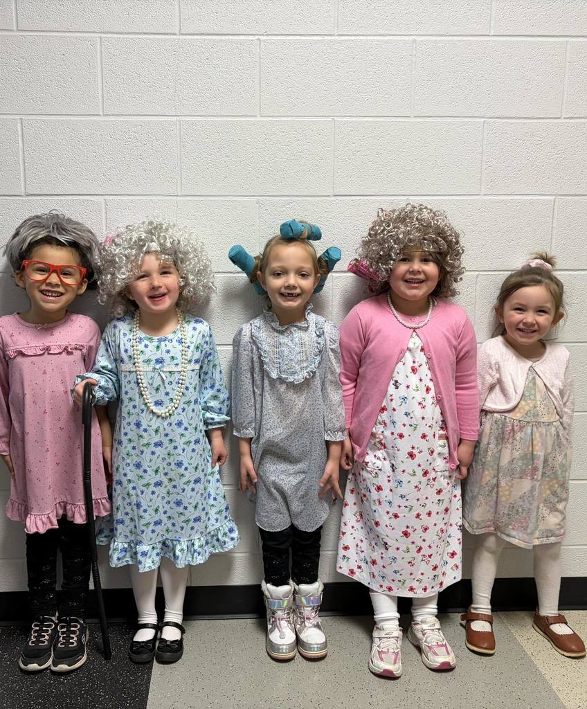 Five young girls pose in a row against a white wall, dressed as "grandmas." Their outfits feature floral nightgowns, cardigans, pearls, and various grey wigs or hair rollers. One child holds a black cane, and they are all smiling brightly for the camera.