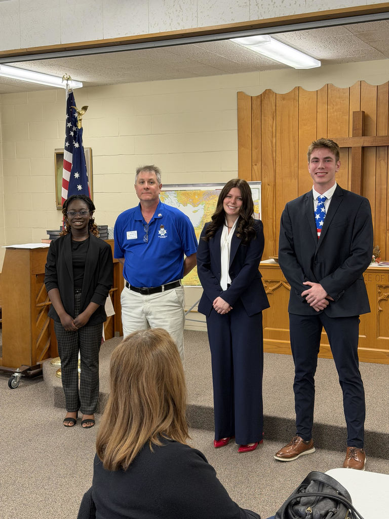 Four individuals stand at the front of a room that resembles a small courtroom or meeting space, facing an audience. Three students in business attire—two women and one man—stand beside an older man wearing a blue polo shirt. An American flag is positioned behind them, along with a wooden wall featuring a cross and a large map. In the foreground, the back of a seated audience member’s head is visible, suggesting a presentation or formal event is taking place.