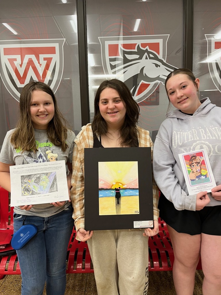 Three female students holding up colorful artwork in front of the Middle School Office