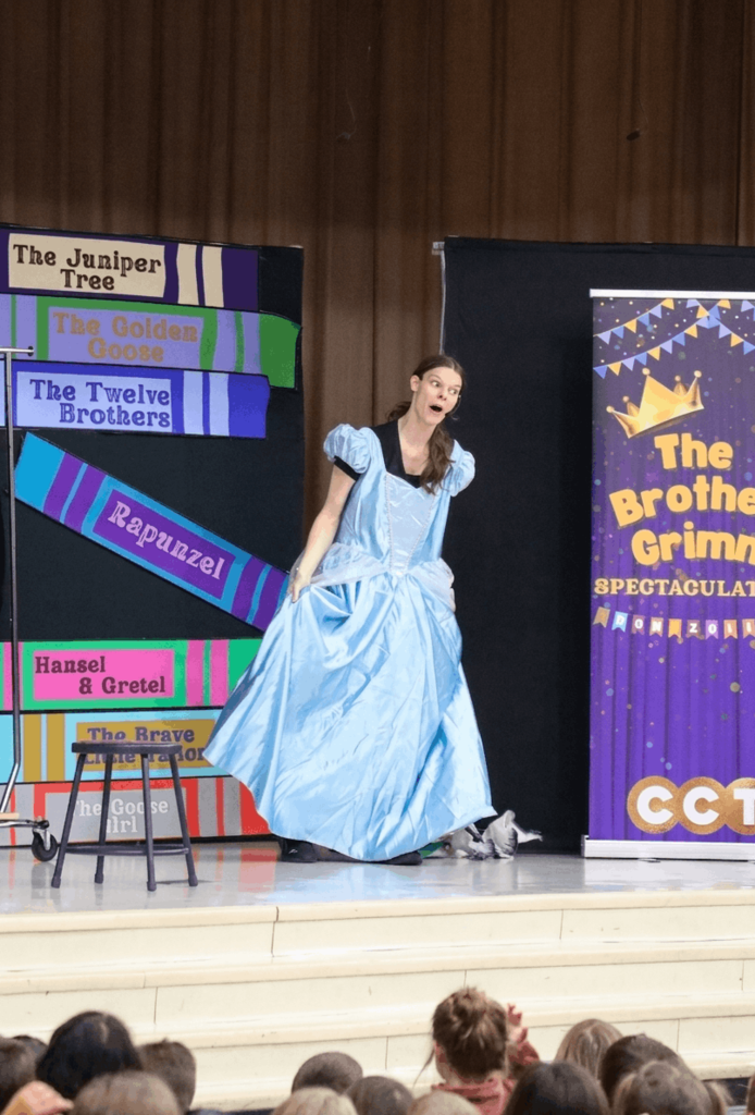 A wide-eyed performer in a shimmering light blue Cinderella-style ballgown stands on a stage, lifting the skirt slightly with an exaggerated expression. Behind them is a set decorated with giant colorful book spines and a purple banner for the Columbus Children's Theatre, with the tops of an audience's heads visible at the bottom of the frame.